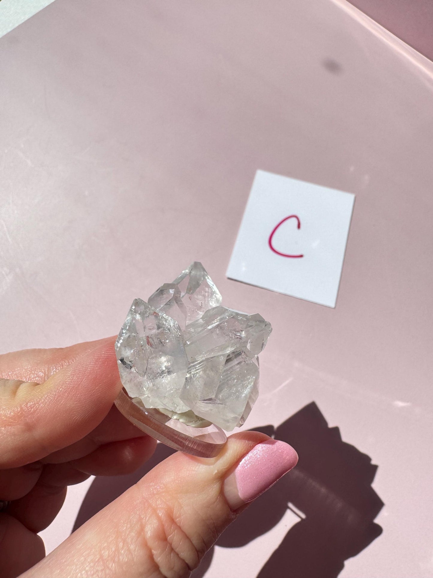A hand with pink nail polish holds an Apophyllite Cluster from India (ITEM C), recognized for spiritual clarity, above a pink surface with a white card marked "C" in red.