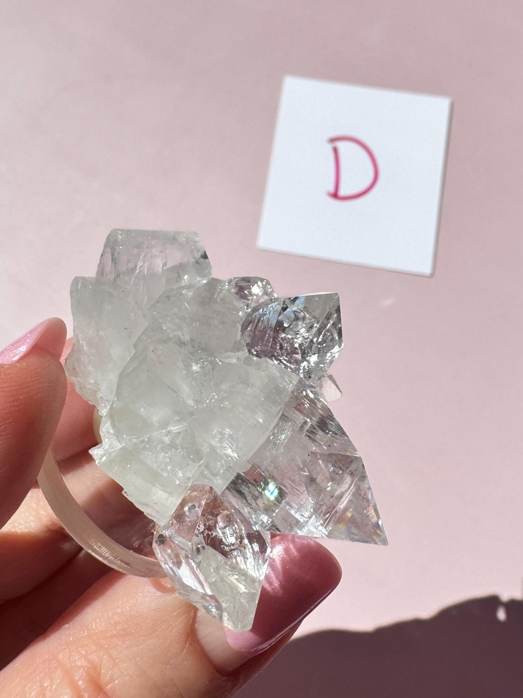 A hand holds the Apophyllite Cluster from India - ITEM D, a quality crystal formation, in front of a pink surface with a red “D” card—ideal for those seeking spiritual clarity and healing crystals.