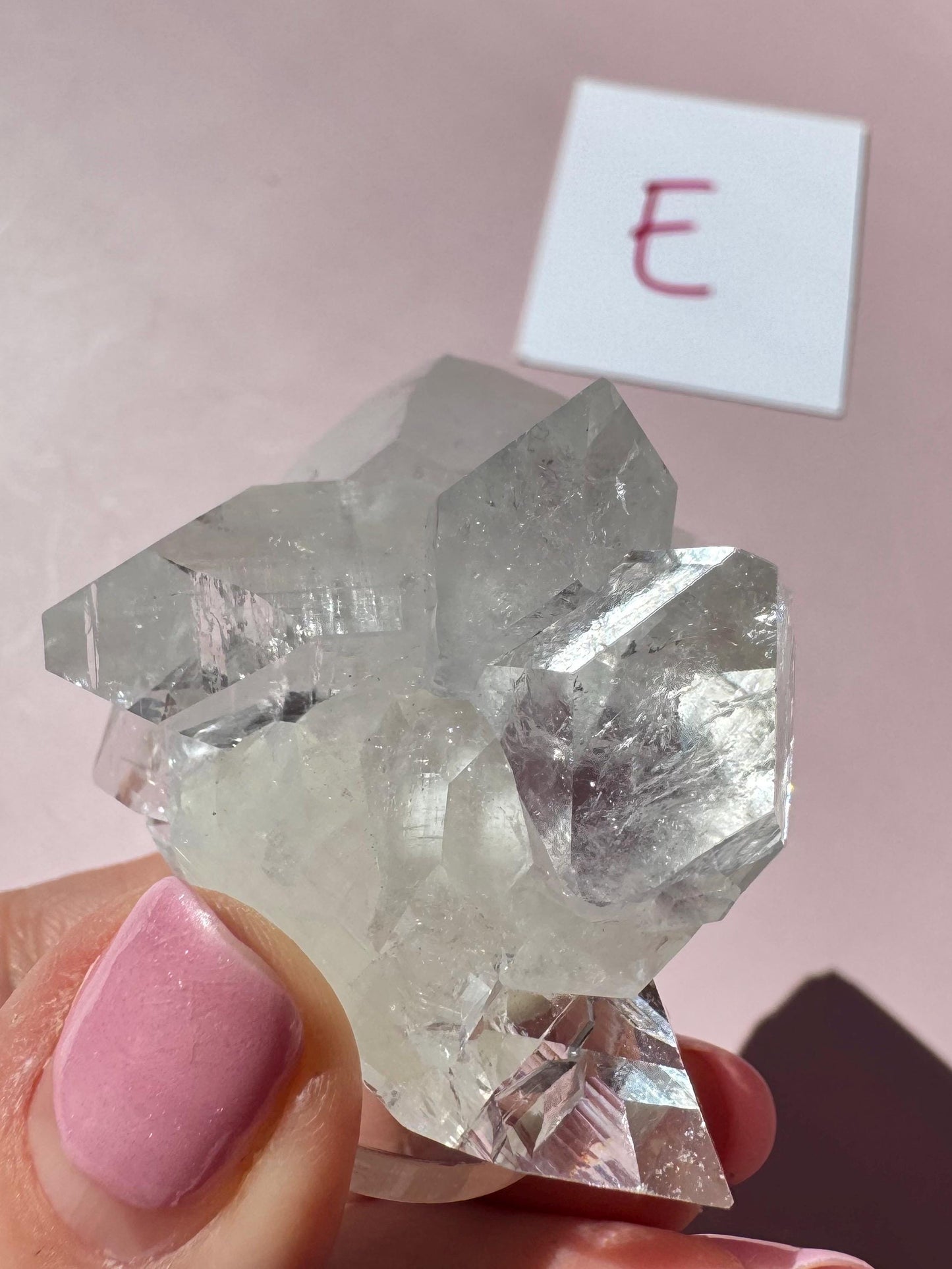 A hand with pink nail polish holds the Apophyllite Cluster from India (ITEM E, nice quality crystal formation), evoking spiritual clarity. A white card marked "E" lies on a pink surface in the background.