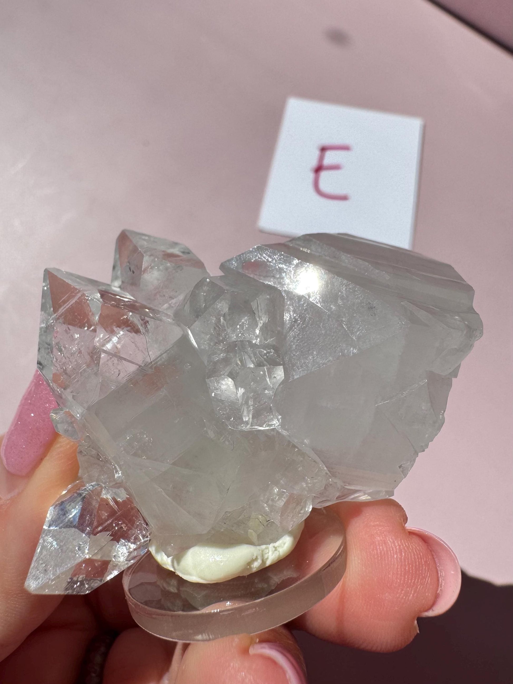 A hand holds the Apophyllite Cluster from India - ITEM E, featuring a nice quality crystal formation on a round base with a white card showing a pink "E"—ideal for those seeking healing crystals and spiritual clarity.