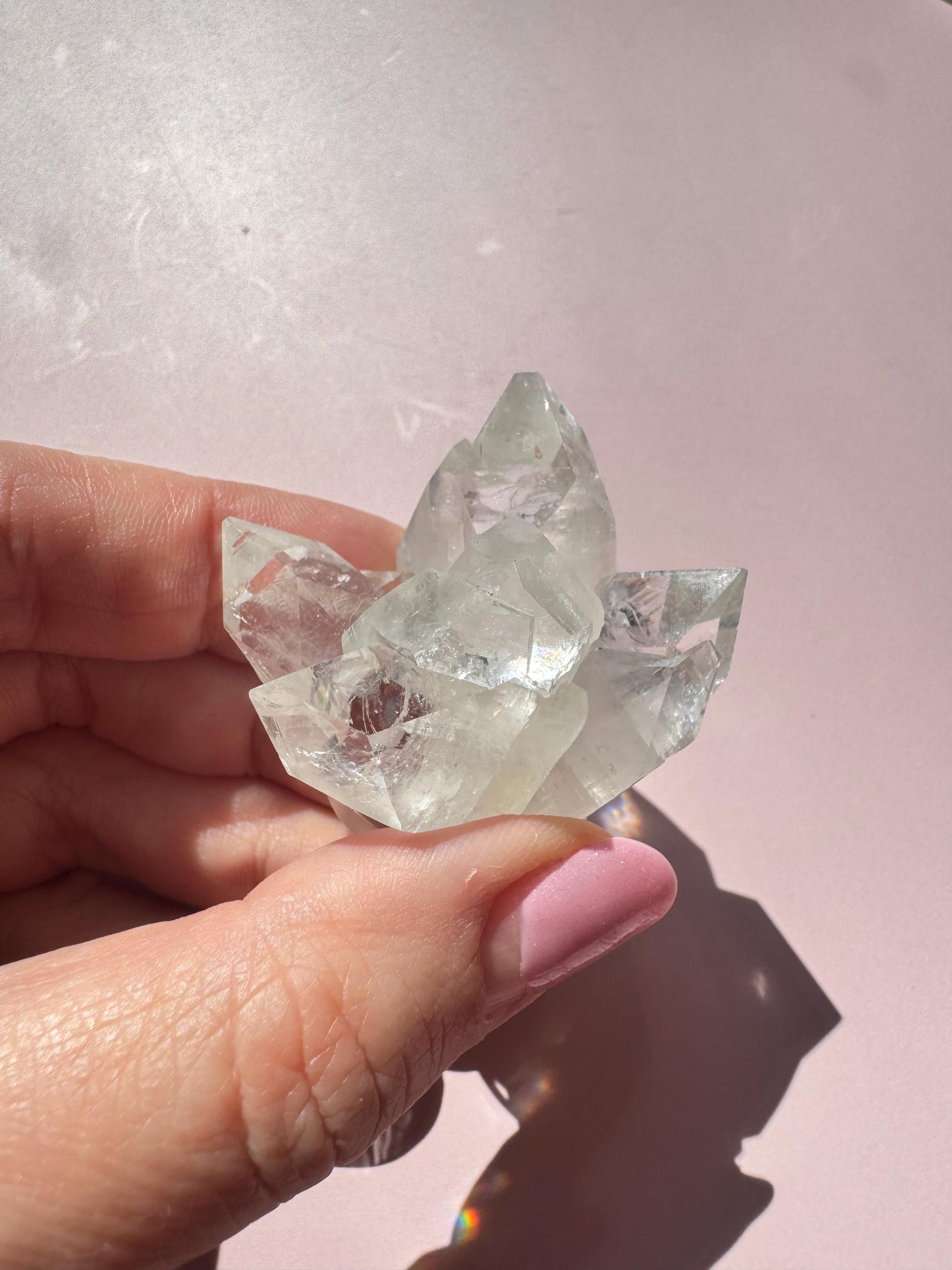 A hand with pink-painted nails holds the Apophyllite Cluster from India - ITEM C, radiating spiritual clarity against a pale pink background.