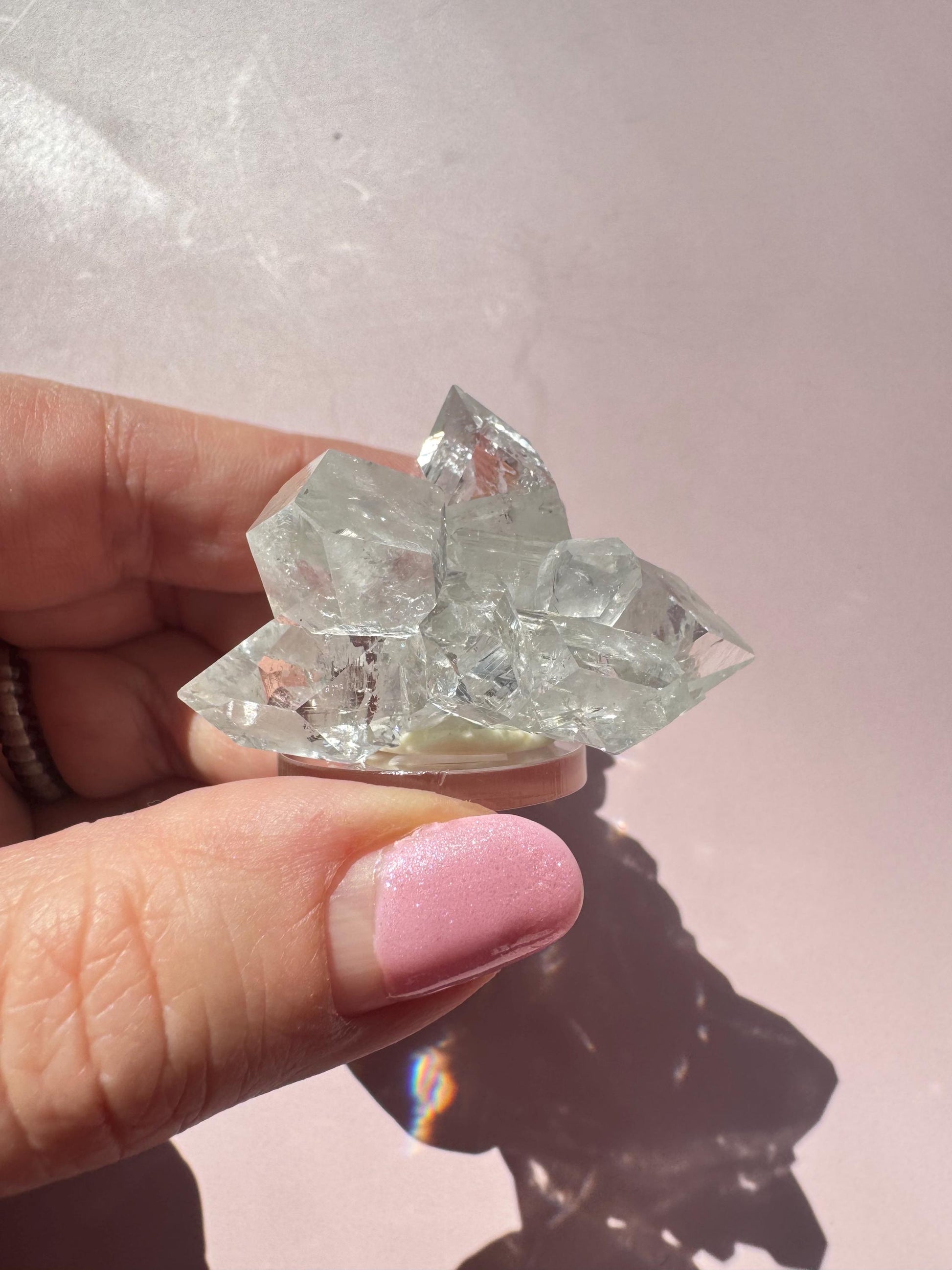 A hand with pink-painted nails holds the Apophyllite Cluster from India (ITEM D), a nice quality crystal formation, against a pale pink background, evoking spiritual clarity and tranquility.