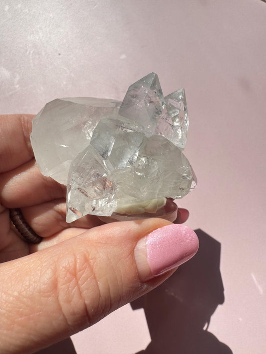 A hand with pink-painted nails holds the Apophyllite Cluster from India (ITEM E), a nice quality crystal formation, against a pale pink background.