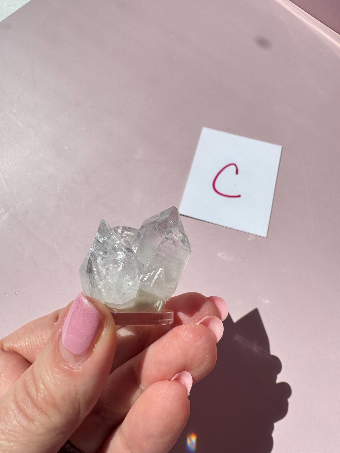 A hand with pink-painted nails holds an Apophyllite Cluster from India (ITEM C), prized for enhancing spiritual clarity; in the background, a white card with a red "C" is visible.