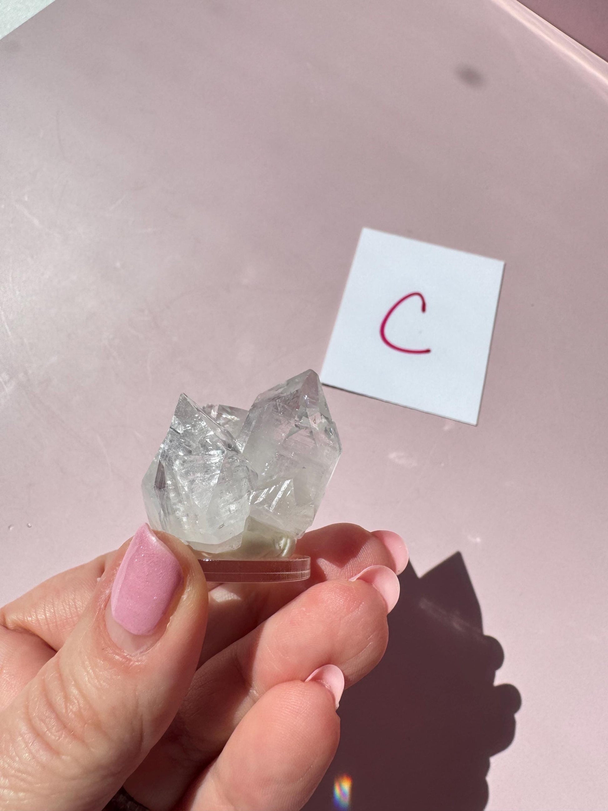 A hand with pink-painted nails holds an Apophyllite Cluster from India (ITEM C), prized for enhancing spiritual clarity; in the background, a white card with a red "C" is visible.