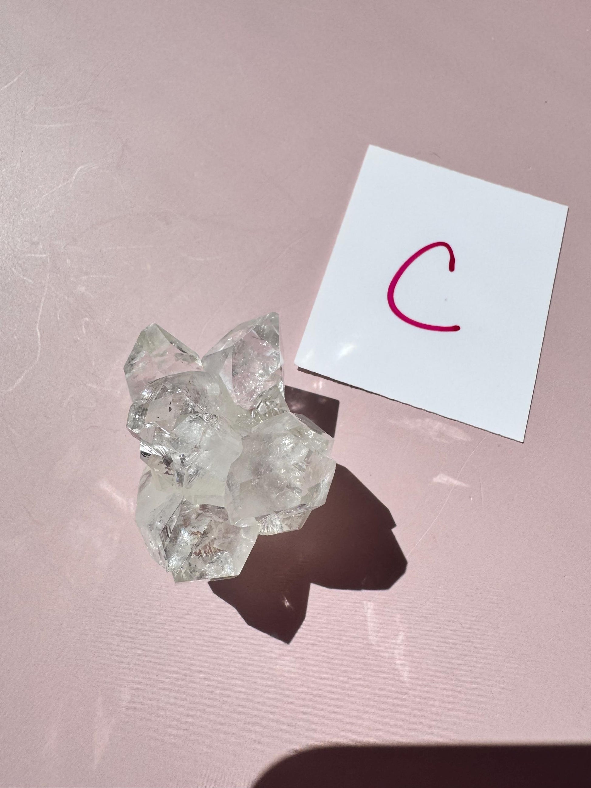 An Apophyllite Cluster from India (ITEM C) rests beside a white card with a red handwritten "C" on a pink surface, evoking spiritual clarity.