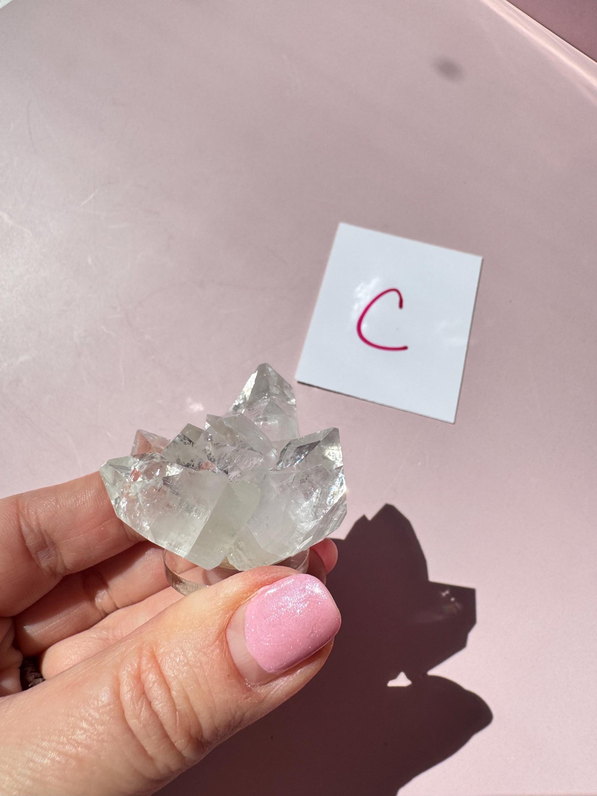 A hand with pink nail polish holds the Apophyllite Cluster from India - ITEM C near a white card with a pink "C" on a pale pink surface, evoking spiritual clarity.