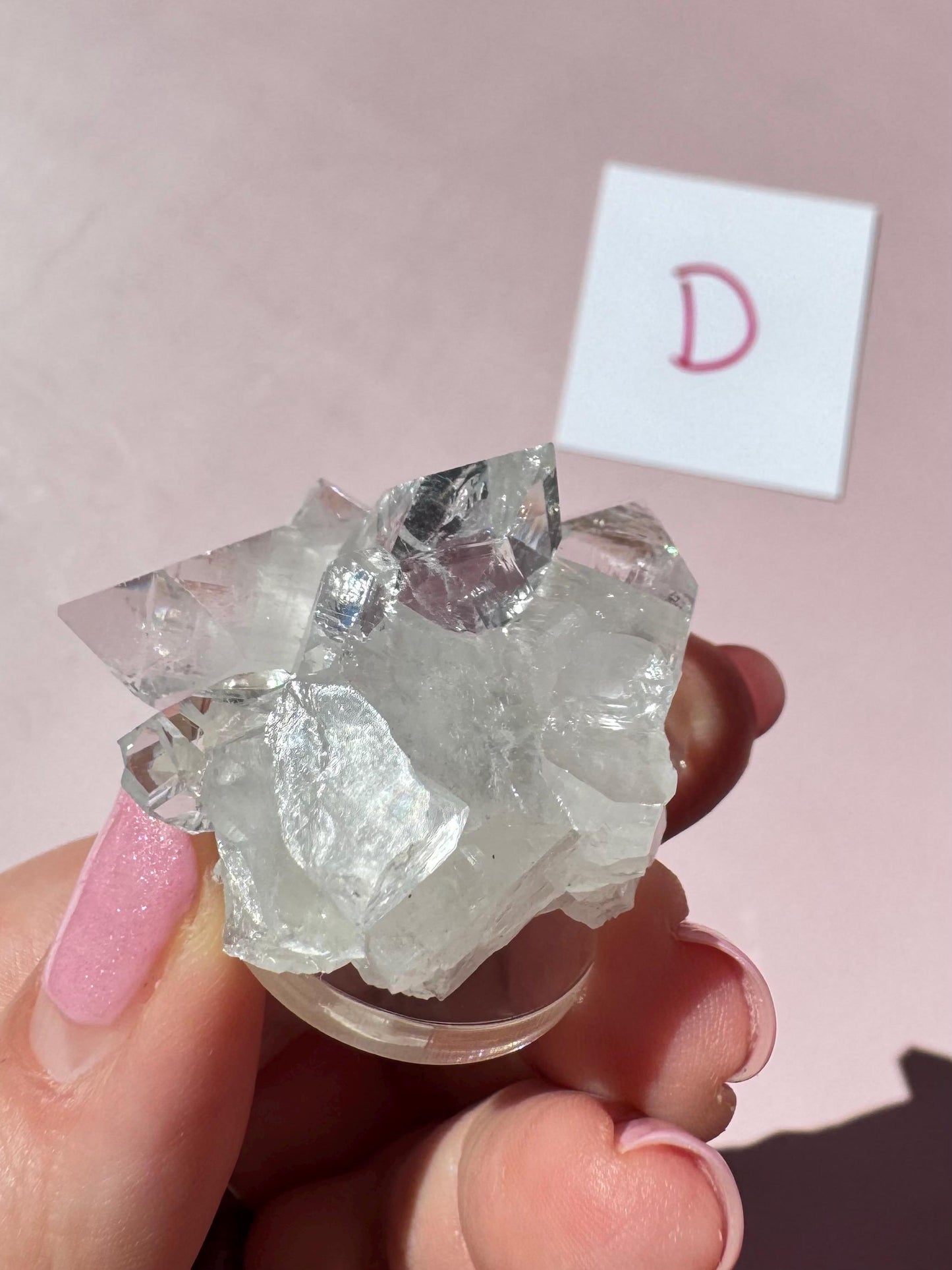A hand with pink nails holds the Apophyllite Cluster from India - ITEM D (nice quality crystal formation); a white card with a pink "D" sits in the background on a pink surface, showcasing this beautiful healing crystal.