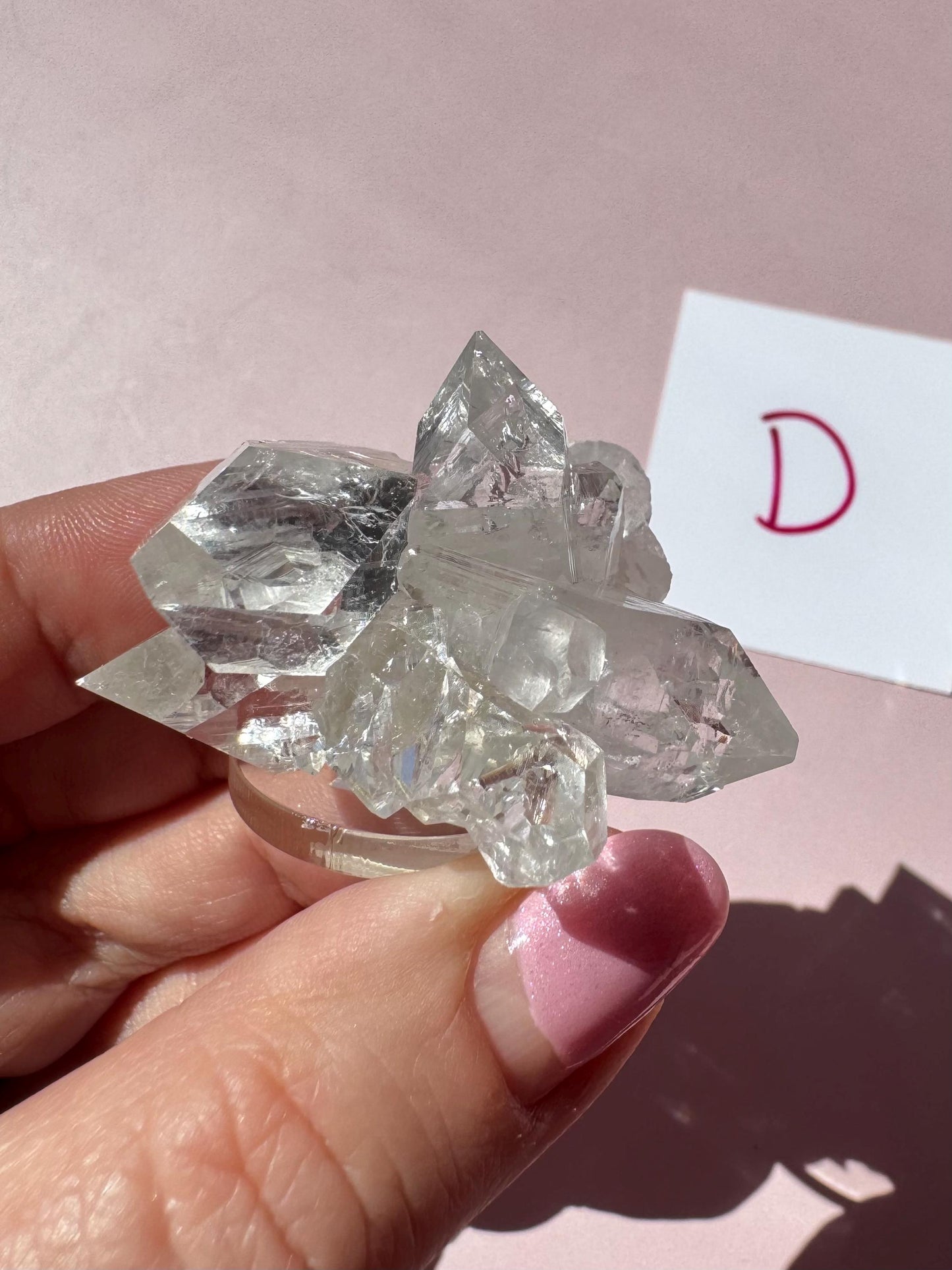 A hand with pink nail polish holds the Apophyllite Cluster from India (ITEM D, nice quality crystal formation); a card labeled "D" appears in the background.
