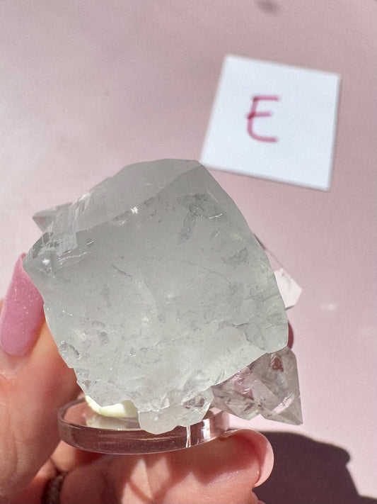 A hand holds an Apophyllite Cluster from India (ITEM E, nice quality crystal formation); behind it, a white card with a red "E" rests on a pink surface, evoking spiritual clarity and the healing properties of this crystal.
