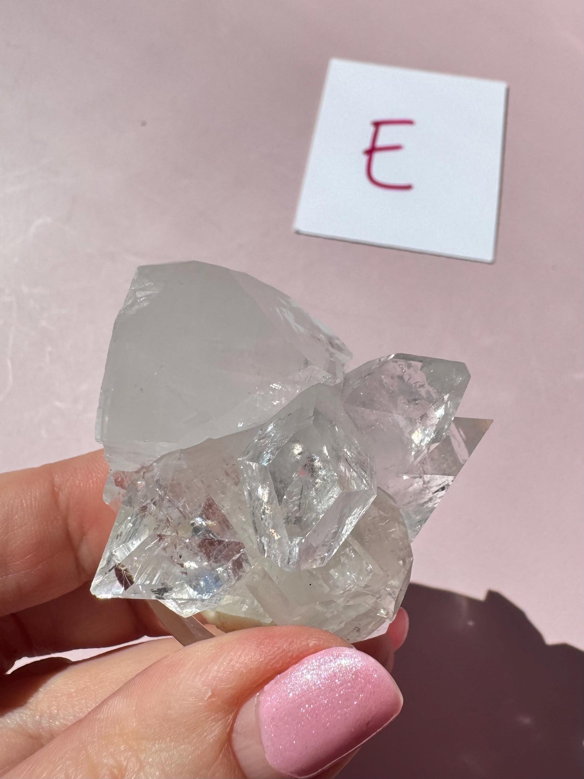 A hand with pink nail polish holds an Apophyllite Cluster from India (ITEM E – Nice Quality Crystal Formation); in the background, a white card with a red "E" sits on a pink surface, enhancing the crystal’s spiritual vibe.