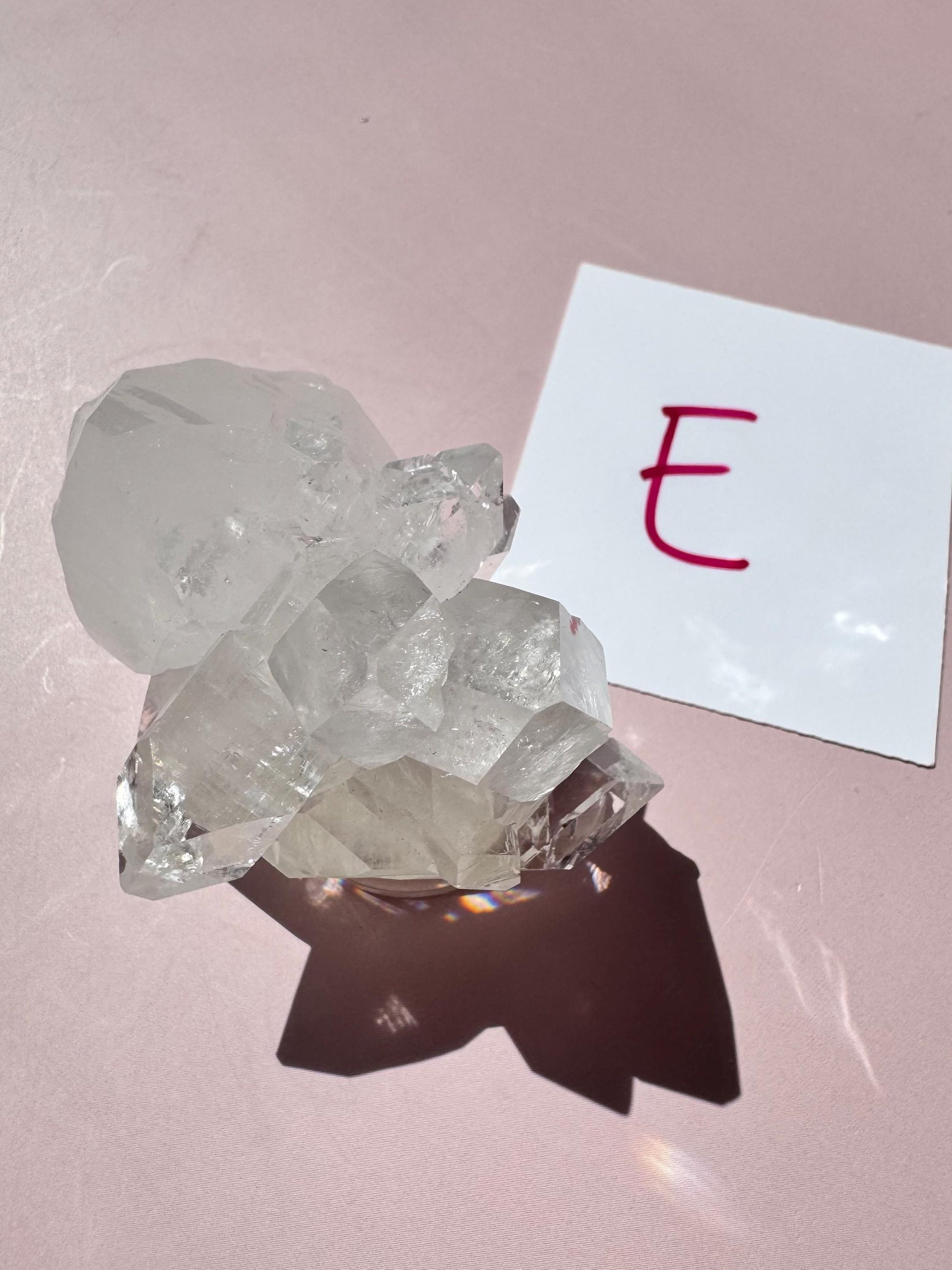 A nice quality Apophyllite Cluster from India (ITEM E) rests on a pink surface beside a white card with a red "E," radiating spiritual clarity and positive energy.