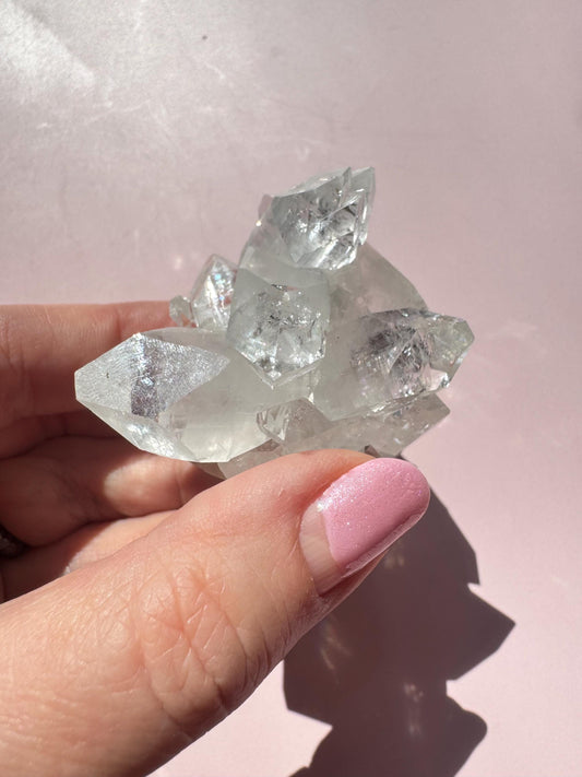 A hand with pink nail polish holds the Apophyllite Cluster from India - ITEM F, a nice quality crystal formation, against a light pink background.
