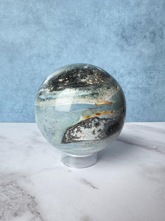 A Light Blue and Dark Green Sea Jasper Sphere (ITEM D) from Madagascar, 74.2 mm in diameter, sits on a clear stand against a blue and white background—a sophisticated piece for any healing crystals collection.